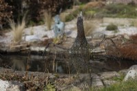 A cormorant sculpture made of recycled wire and mesh in front of image beside a pond, with Marissa, a copper bronze sculpture by Jenny Wynne Jones, sat amongst gravel and grasses in background. Selective focus. February. 