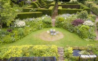 View over wide border of Alchemilla mollis and mixed border surrounding lawn with metal table and chairs painted pastel colours and formal hedges of Yew in Background. Image taken from drone. July.