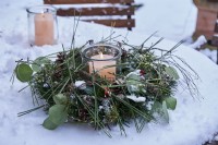 A pillar candle in storm glass surrounded by wreath decorated with pine needles and branches and Eucalyptus foliage on a snowed table