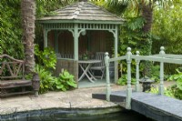 Painted Gazebo in poolside setting with surrounding planting of Bamboo, Palms, Hostas and Fatsia japonica and bridge over pool - Open Gardens Day, East Bergholt, Suffolk 