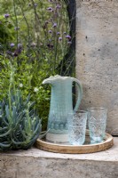 A pale blue jug and two glasses on a tray on the garden wall. Surrouned by: Senecio mandraliscae and Verbena bonariensis growing by the wall. The Shifting Garden, Designers: The Chelsea Gardener.