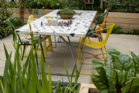 A community table that incorporates two chess boards and is place for people to come together to eat or play games. Plants in the foreground, red chard and Iris pseudacorus with a terra cotta bowl of Thymus vulgaris. The London Square Community Garden, Gold winner - Chelsea 2023 Designed by: James Smith