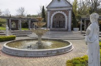 The entrance to the show gardens, with fountain, large, wisteria laden pergola and statues. A duck is swimming on the fountain pool. A classical stone statue is on the right. Brick paving surrounds the fountain pool. Trago Mills show gardens, Devon, UK. May. Spring