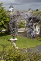 An armillary sundial on pedestal with wisteria covered pergola behind. Trago Mills show gardens, Devon, UK. May. Spring