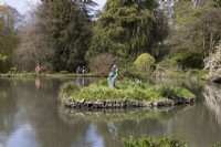 A large lake with an island in the middle and trees in the background. The island has a sculpture of a young girl swinging a small child into the air. Marwood Hill Gardens, Devon. Spring. May
