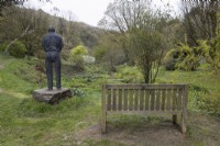 A wooden bench is beside a statue of a man looking out at a view of a bog garden in a valley with various established trees in the background. Marwood Hill gardens. Spring. May. 