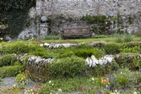A wooden bench sits at the base of an old stone wall, with the Scree Beds in front and pedestals beside the bench with peacock statues on them. Spring. May. Marwood Hill gardens. Devon.