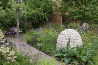 A path leads to a tranquil seating space in the shade of Betula nigra, passing through herbaceous borders of perennials, flanked by a stone cairn.