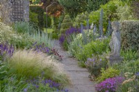 View of mixed perennials borders either side of a stone path in an informal country cottage garden in Summer - June
