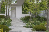 A paved stone path leads to a concrete pavilion, through an avenue of multi-stemmed Gleditsia triacanthos, with borders of yew cushions,  grasses, euphorbia and white foxgloves, roses and white laceflower.