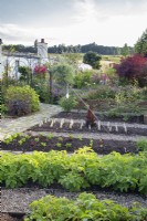 View over vegetable beds divided by gravel paths, beyond crazy paving path and flower beds 