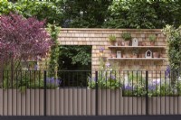 A balcony design, the wall clad in cedar roof shingles and shelves of pots and herbs. The planting is designed to attract pollinators, in planters made from recycled bottles. Design Christina Cobb, Chelsea Flower Show 2023.