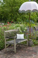 Rustic wooden seat on brick flooring with parasol, fronting lush border of perennial plants - Open Gardens Day, Tuddenham, Suffolk