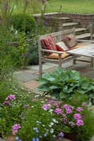 Sunken patio with table, seat and cushions and raised borders of perennial plants. Steps lead up to lawn at higher level - Open Gardens Day, Tuddenham, Suffolk



