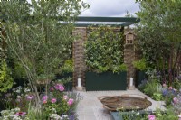 A formal courtyard is enclosed in willow and hazel fencing screens, supporting fragrant star jasmine, Trachelospermum jasminoides, which grows in tall planters with herbs. A water feature is crafted from reclaimed aluminium. Planted in the beds are hawthorn and Tibetan cherry trees, Rosa 'Princess Alexandra of Kent' and herbaceous perennials.