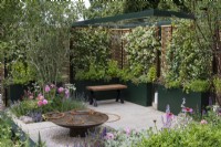 A formal courtyard is enclosed in willow and hazel fencing screens, supporting fragrant star jasmine, Trachelospermum jasminoides, which grows in tall planters with herbs. A water feature is crafted from reclaimed aluminium. Planted in the beds are a hawthorn tree, roses and herbaceous perennials.