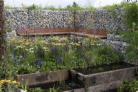 A wildlife friendly, sustainable city space with natural stone gabion walls and wooden bench, reclaimed timber pools, and borders of grasses and nectar rich flowers.