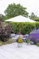 Parasol in weighted container planted with Gaillardia, Kniphofia, Agapanthus and Agastache next to table and chairs on porcelain patio