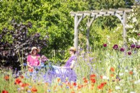 Women chatting sat at table next to wooden arch in garden beyond flowers