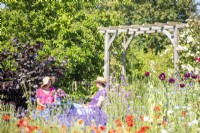 Women chatting sat at table next to wooden arch in garden beyond flowers