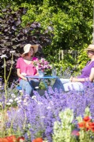 Women chatting sat at table in garden beyond flowers