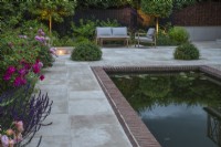Garden lighting on sandstone terrace with rectangular pool and furniture, Pittosporum topiary balls for light concealment and borders of Roses 