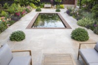View down on rectangular brick lined pool on sandstone terrace with furniture and borders of David Austin roses; Lavender; Ballota; Salvia 'Caradonna' and Pittosporum topiary balls