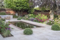 Brick edged rectangular pool on terrace of sandstone paving with garden furniture.  Borders include David Austin roses; Salvia nemorosa 'Caradonna', lavender, Ballota and Pittosporum topiary balls