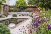 Seating area on sandstone terrace with brick edged rectangular pool and border of David Austin roses, 