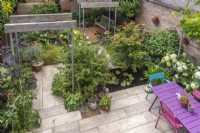 Birds eye view down on small enclosed paved urban garden featuring patio, Industrial style single pergolas, containers, rectangular pool with waterlilies, colourful table and chairs. Planting inc: Hydrangea 'Annabelle', Acers, Wisteria and Fuschias