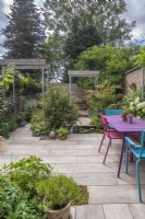 Small paved urban garden featuring industrial style single pergolas with scaffold poles and half oak sleepers; pool and colourful table and chairs.  Plants inc: Acers, Fuchsias and perennials