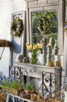 Outdoor arrangement featuring arched mirrors and shelves with potted plants, wreath and bunch of daffodils in glass vase.