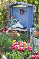 Woman reading a magazine in a covered bench in spring garden with tulips.