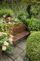 Red oxide painted metal bench surrounded by shrubs and summer flowers in a garden in August.