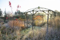 Sunlit wooden rustic gazebo surrounded by ornamental grasses and perennials in frost.