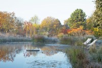 Natural swimming pool with central millstone water feature and seating area with deckchairs surrounded by frosted ornamental grasses 