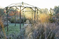 Frosted wooden rustic gazebo and seating area surrounded by ornamental grasses at sunrise.