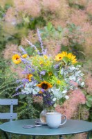 Bouquet containing Helianthus - Sunflowers, Eucalyptus sprigs, Tagetes 'Lemon Gem', Perovskia and Calendula 'Indian Prince' in glass vase on small garden table with snips and a mug