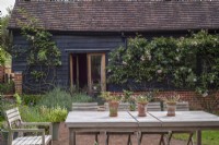 Table and chairs with containers of Erigeron karvinskianus on brick paved terrace in front of barn with Rosa 'The Generous Gardener'