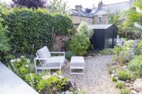 Small garden with black shed and curved gravel path running through the middle towards a seating area with Chair and footstool in the foreground