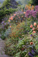 Paved path leading through misty autumnal borders in cottage-style garden, tiled summerhouse behind