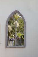 An arched, stone edged mirror on a cream wall reflecting a metal pot with pansies, cordyline australis and a variety of other shrubs and plants. June. 