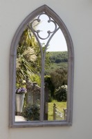 An arched gothic style mirror on a cream wall reflects a balcony, with Cordyline australis and far reaching countryside view. 