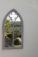 An arched gothic style mirror on a cream wall reflects a balcony, with Cordyline australis and far reaching countryside view. May. 