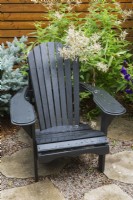 Black wooden Adirondack chair on flagstone and pebble patio bordered by Picea glauca - Blue Colorado Spruce Tree and Persicaria wallichii - Fleeceflower shrub in backyard in summer.