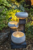 Illuminated cascading granite bowls water fountain in mulch border in backyard garden at dusk in summer.