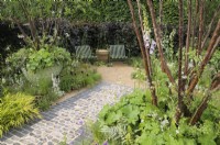 An urban garden with a straight path made of brick and flint leading to a small seating area with two green HAY Palissade Lounge Chairs enclosed by copper beech hedging.  Multi-stemmed Prunus serrula trees - Tibetan cherry - in large planters with Digitalis purpurea 'Dalmatian White', Alchemilla mollis and Athyrium niponicum 'Metallicum'.  The Traditional Townhouse Garden, RHS Hampton Court Palace Garden Festival 2023.  