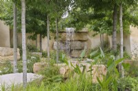 Woodland plants and trees frame a waterfall cascading over slabs of rock providing a dynamic focal point.  America's Wild, presented by Trailfinders  and  Visit The USA, RHS Hampton Court Palace Garden Festival 2023.  