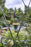 View through a green wrought iron gate of a formal garden based on the historic courtyard gardens of Charleston, South Carolina, with a pineapple water feature representing the famous Charleston waterfront fountain.  Explore Charleston - Welcome to Charleston, RHS Hampton Court Palace Garden Festival 2023.  