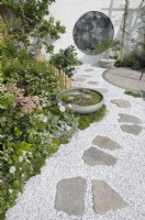 A grey stone bowl with shallow water and aquatics on the edge of a winding gravel path with irregular stone slabs leading to a round mottled antique mirror on the back wall - reminiscent of an Oriental moon gate.  The Lunar Garden, RHS Hampton Court Palace Garden Festival 2023.  